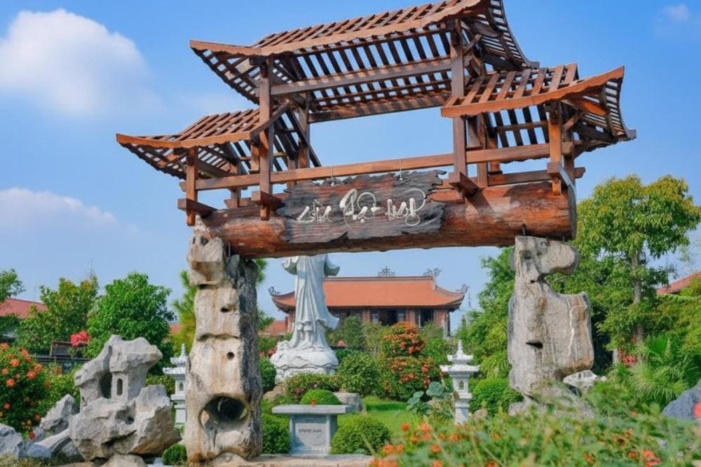 Cổng gỗ ba tầng mái cong 3 tầng kiểu Á Đông, đặt trên hai trụ đá tự nhiên, dẫn vào khu vườn xanh tươi có công trình mái ngói đỏ truyền thống.